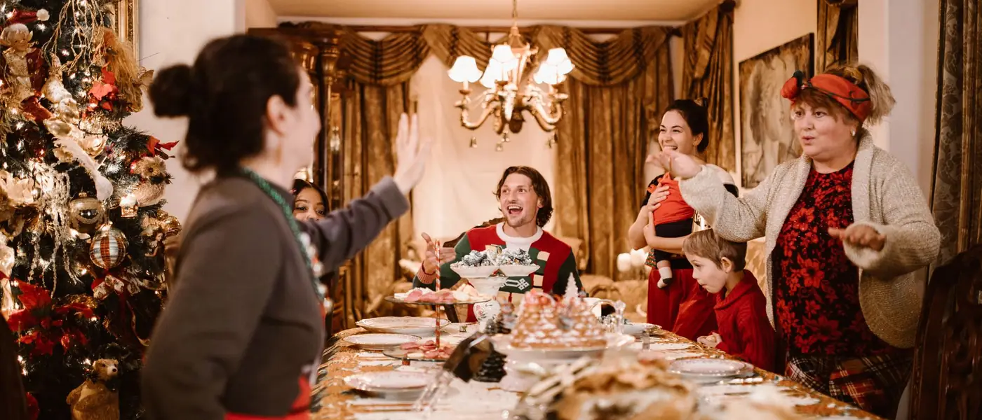 A full house round a long Christmas dinner table, tree lit in the corner, paper hats on